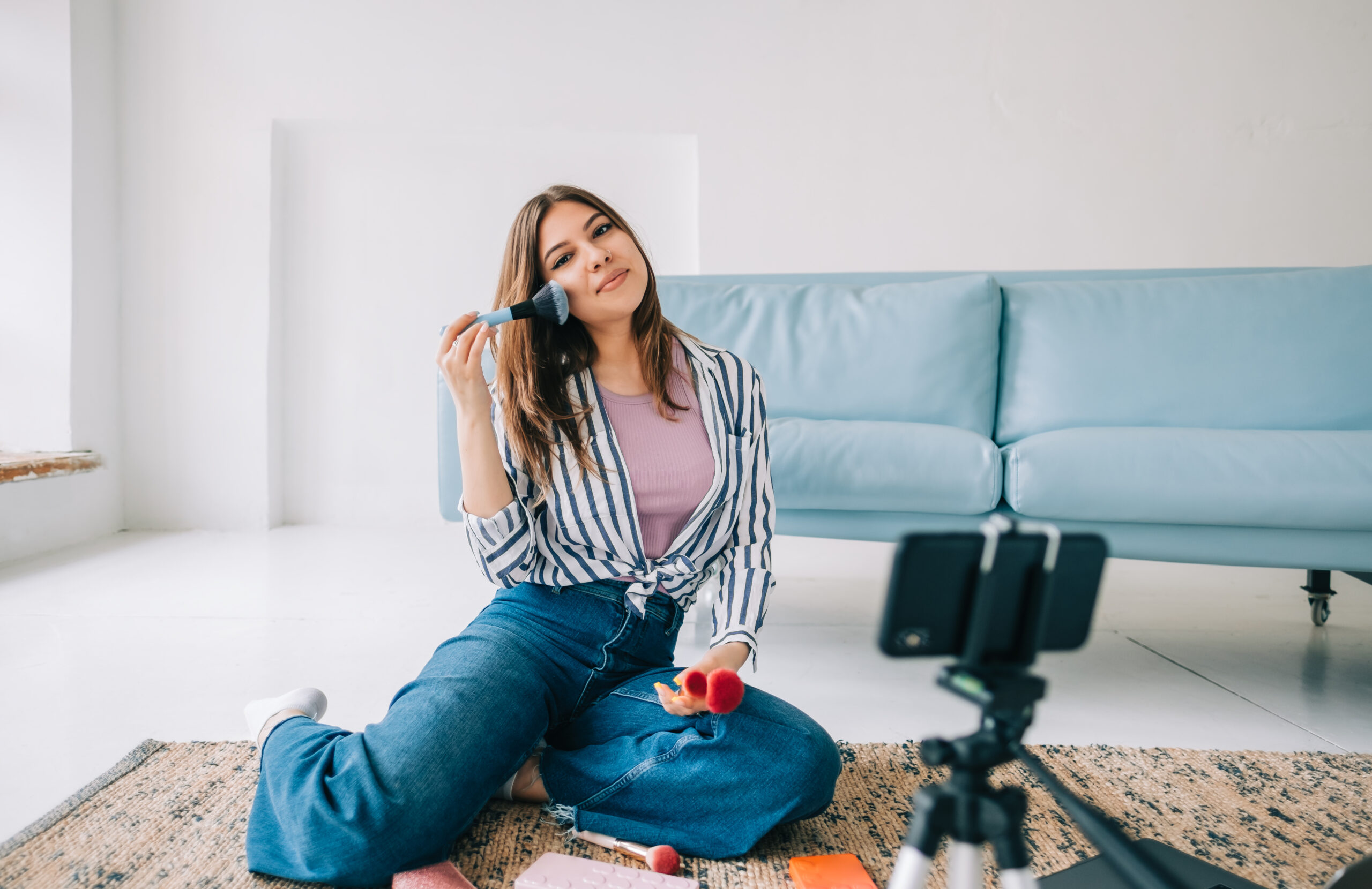 Attractive caucasian young woman video blogger showing beauty products via her blog on social media, holding makeup brushes looking at smartphone camera fixed on tripod.